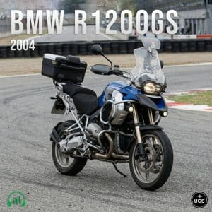 Blue and silver BMW R1200GS 2004 motorcycle on racetrack, with windshield, side mirrors, rear storage box, barriers and stands.